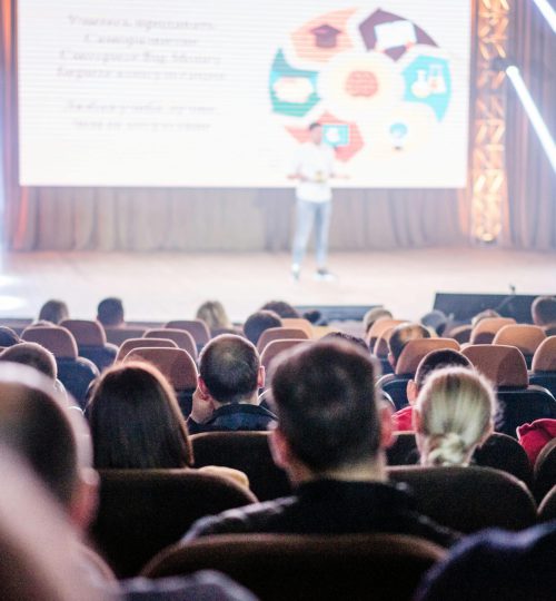 Audience attentively watching a presentation in a conference setting, with stage lights and speaker visible.